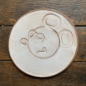 Pale glazed ceramic plate with a face made from impressed letterpress blocks, on wooden floor
