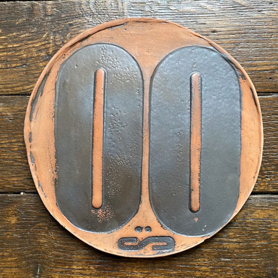 Round, decorative plate with black glaze details and face made from letterpress block impressions on a wooden floor